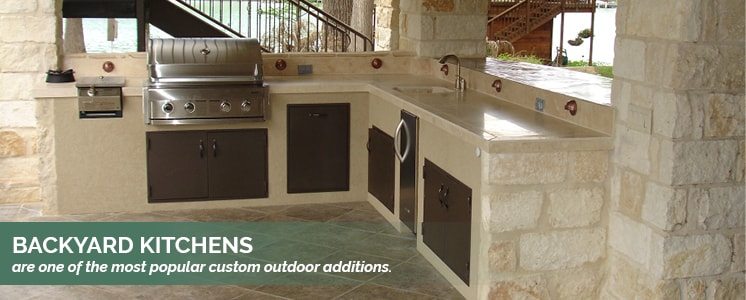 An outdoors tan themed kitchen with pillars of large stone bricks holding an unseen roof casting the whole set-up into shade with a banner of text along the bottom that reads "Backyard kitchens are one of the most popular custom outdoor additions."