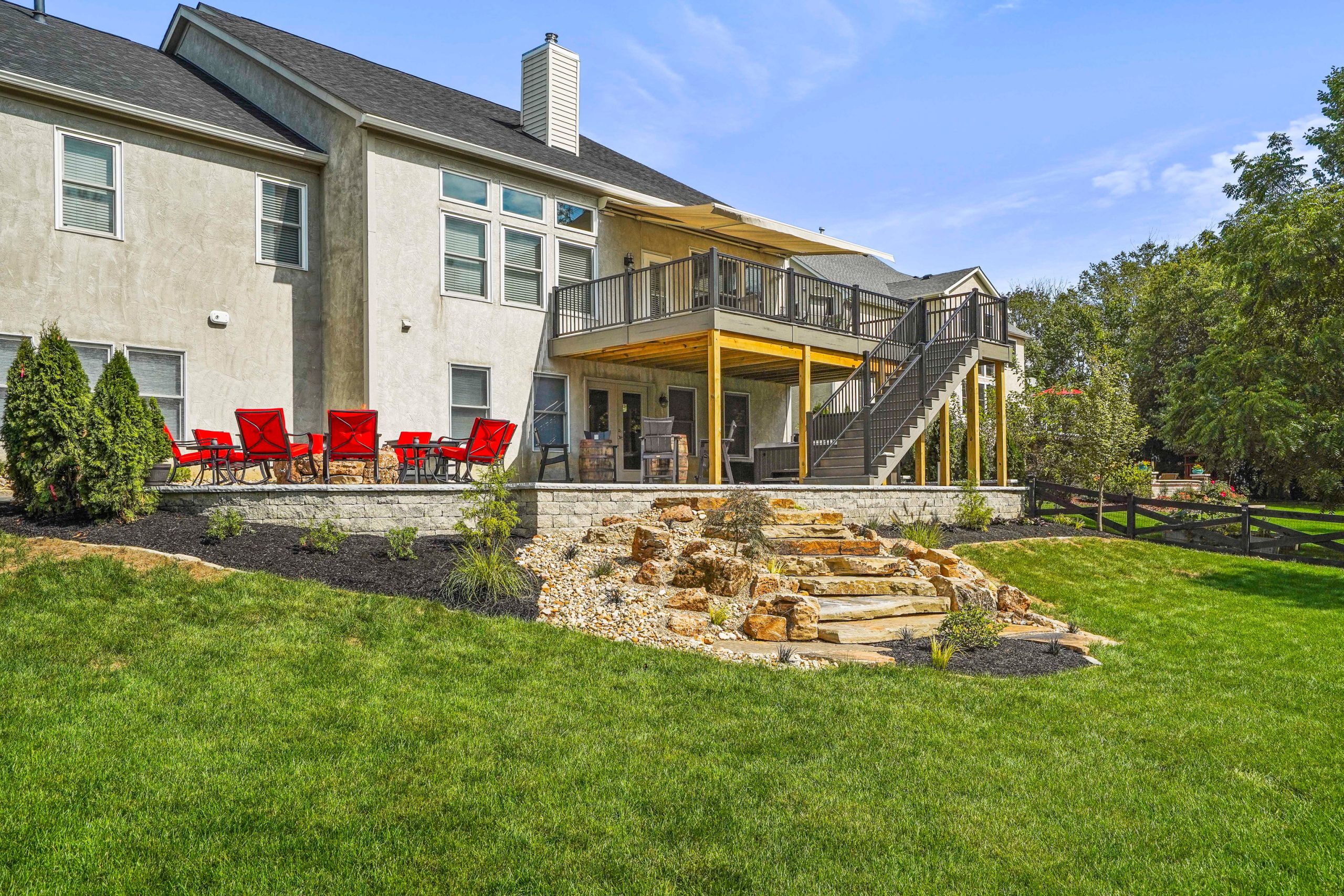 Full view of a storey house with patio on one side and fireplace deck on the other. There is a lawn and shrub around the rock stairs within the house fence. There are trees outside the fence.