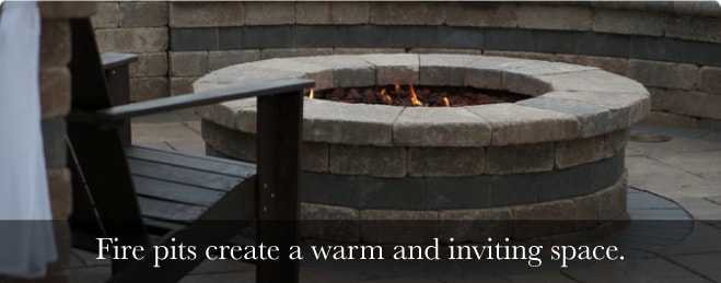 A low close up shot of a round brick firepit with a wooden chair out of focus to the left side with text below reading "Fire pits create a warm and inviting space."