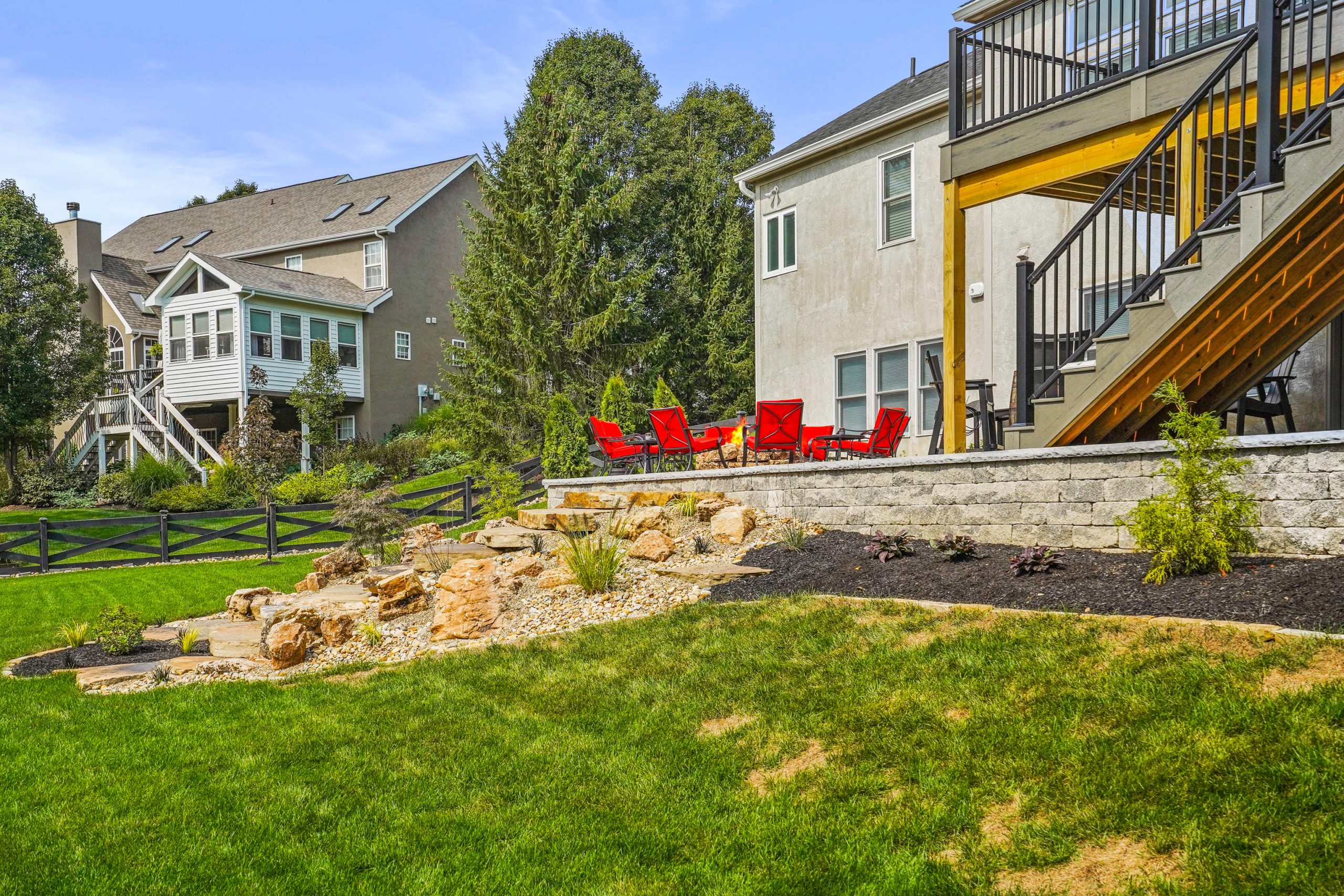 Lawn view of shrubs garden around rock stairs. The rock stairs lead to fireplace deck with red chairs. There are trees and lawn on neighbor's property to the left of fireplace.