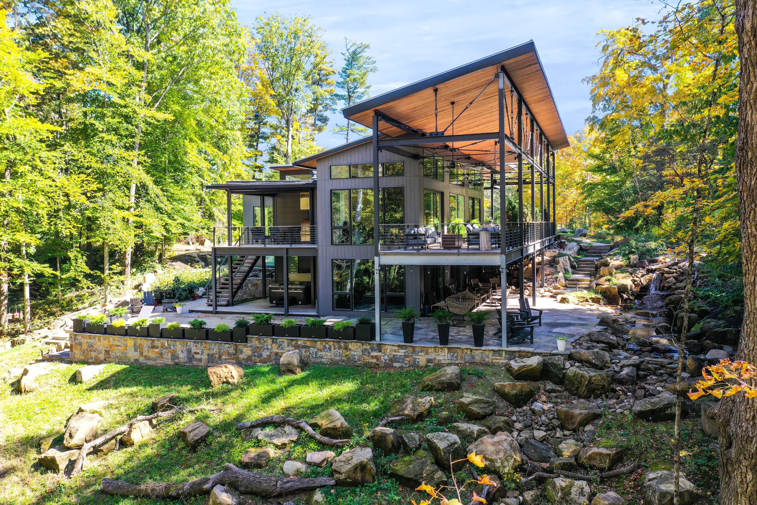 Full view of home with lawn and shrubs on one side. The home is surrounded by colorful forest trees.