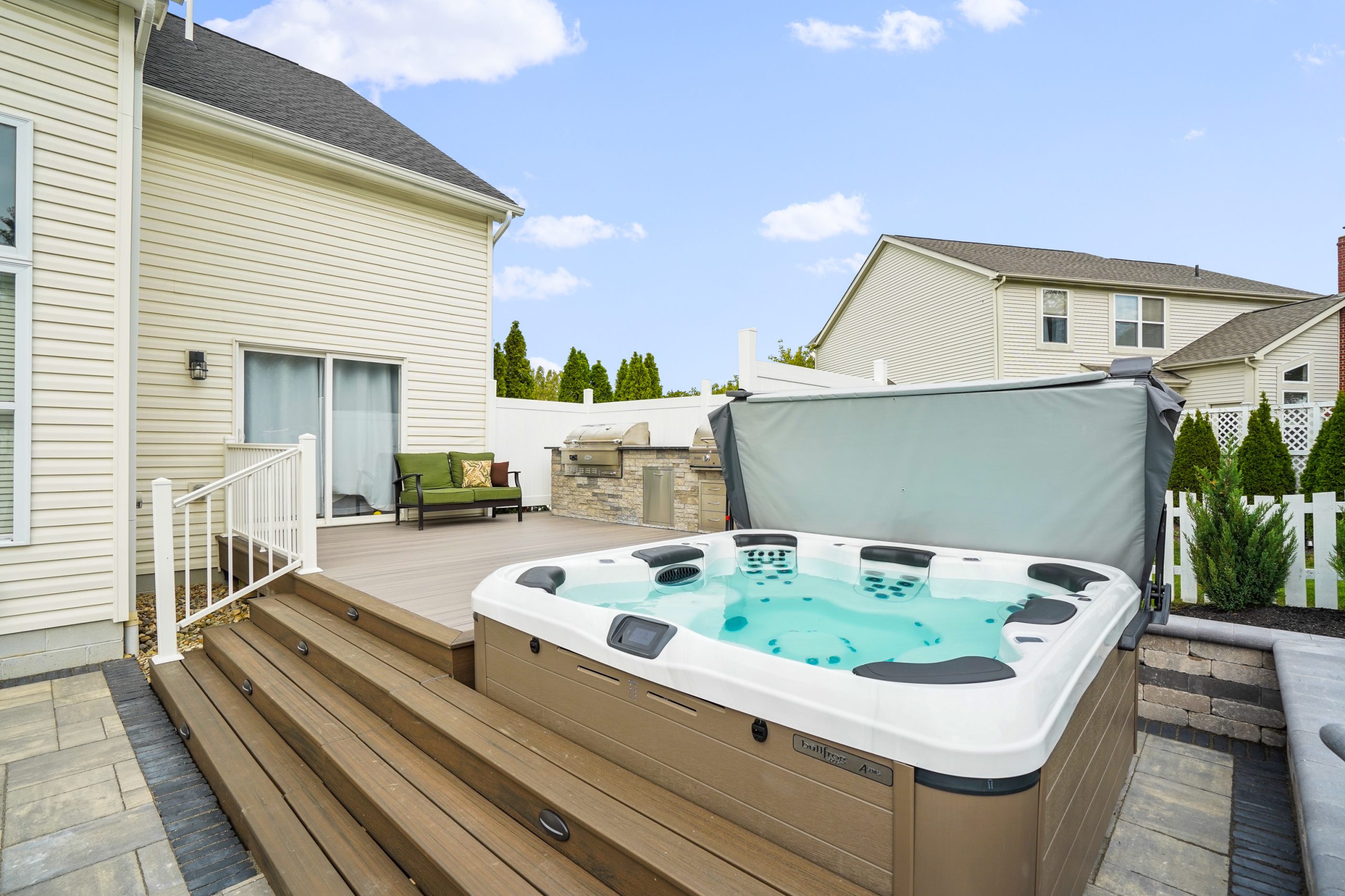 Side-by-side jacuzzi and kitchen on deck with shrubs, trees and neighbor’s house behind.