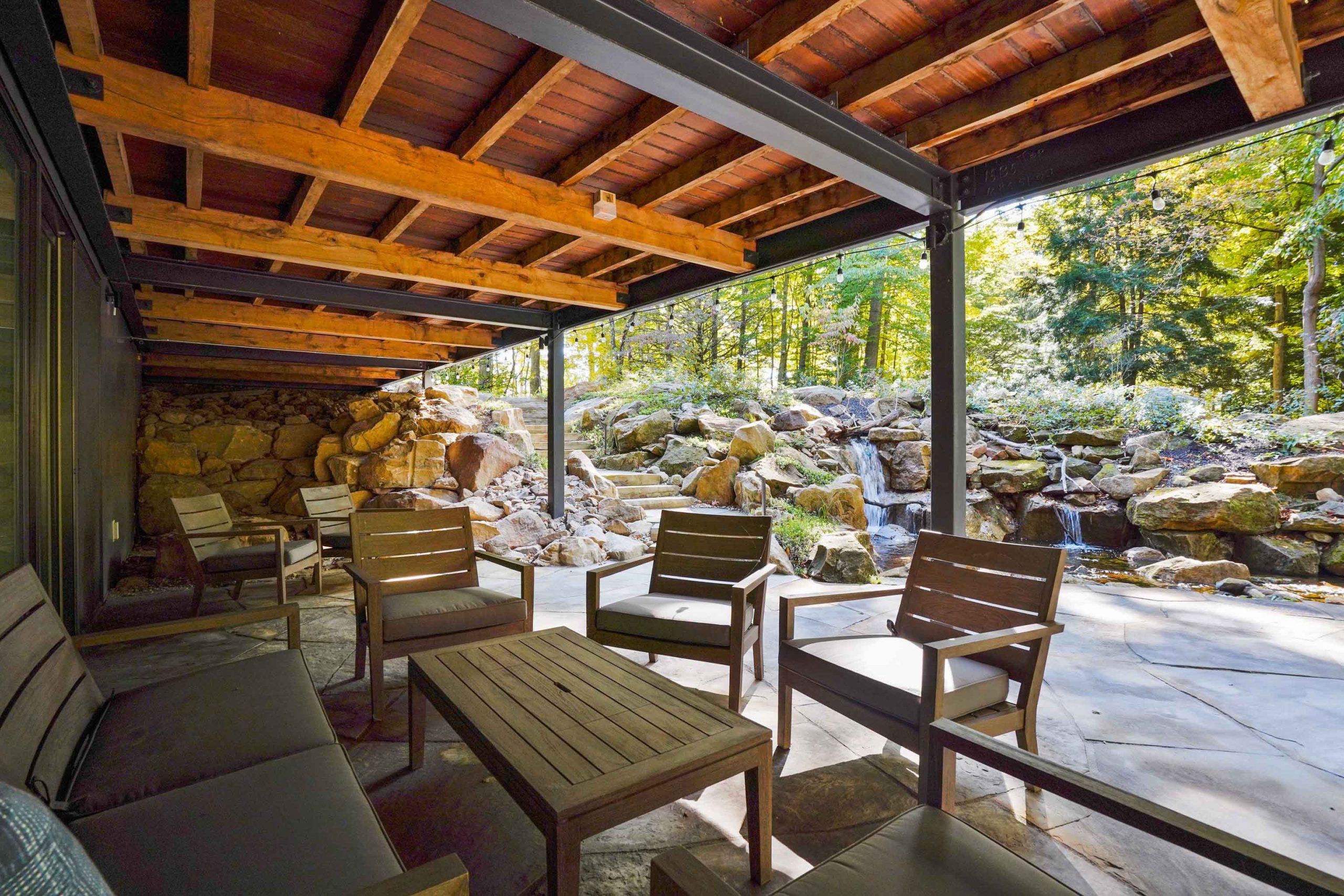 Lounge area under patio overlooking pondless waterfall and colorful tall trees.