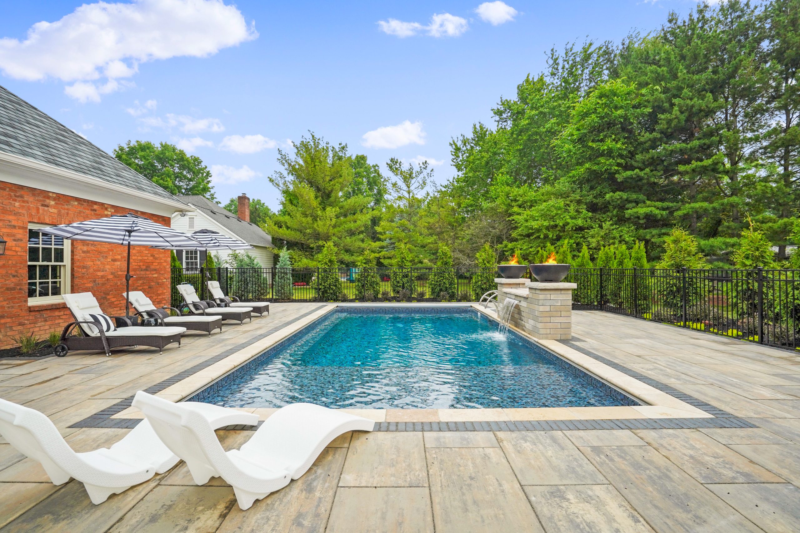 Pool with umbrella chairs within a fenced area overlooks hedges and trees behind the fence.