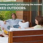 A group of adults sitting in pairs across from each other in a very clean and tidy outdoors patio with cushioned wooden benches smiling at each other with text reading "Whether you’re cooking, entertaining guests or just enjoying the moment, life is more relaxed outdoors."