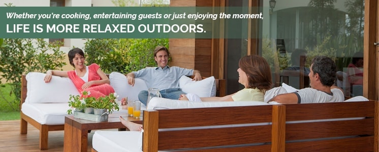 A group of adults sitting in pairs across from each other in a very clean and tidy outdoors patio with cushioned wooden benches smiling at each other with text reading "Whether you’re cooking, entertaining guests or just enjoying the moment, life is more relaxed outdoors."
