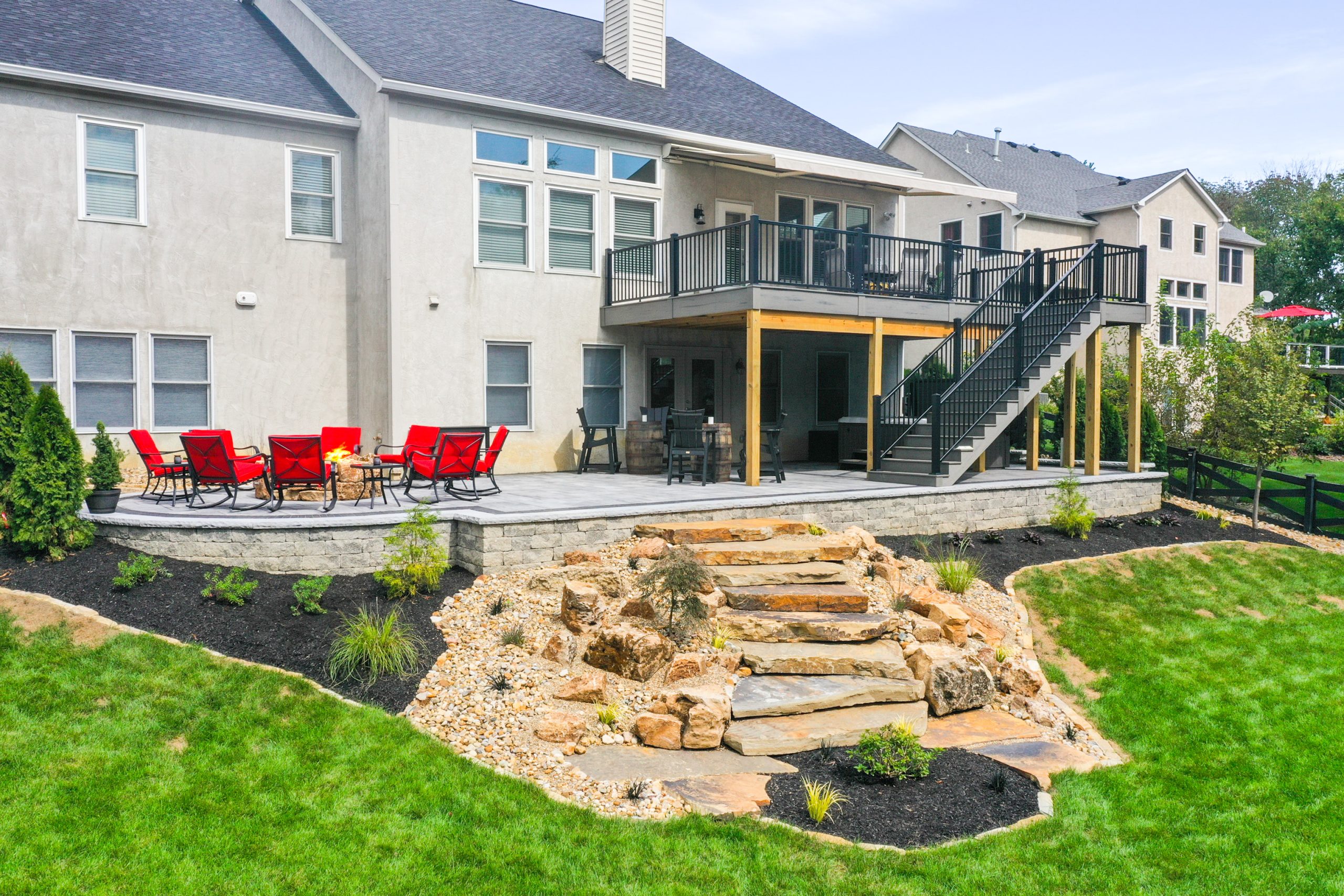 Full view of a storey house with patio upstairs, jacuzzi in shade downstairs, fireplace with red rocking chairs, stairs hardscape surrounded by shrubs and lawn.