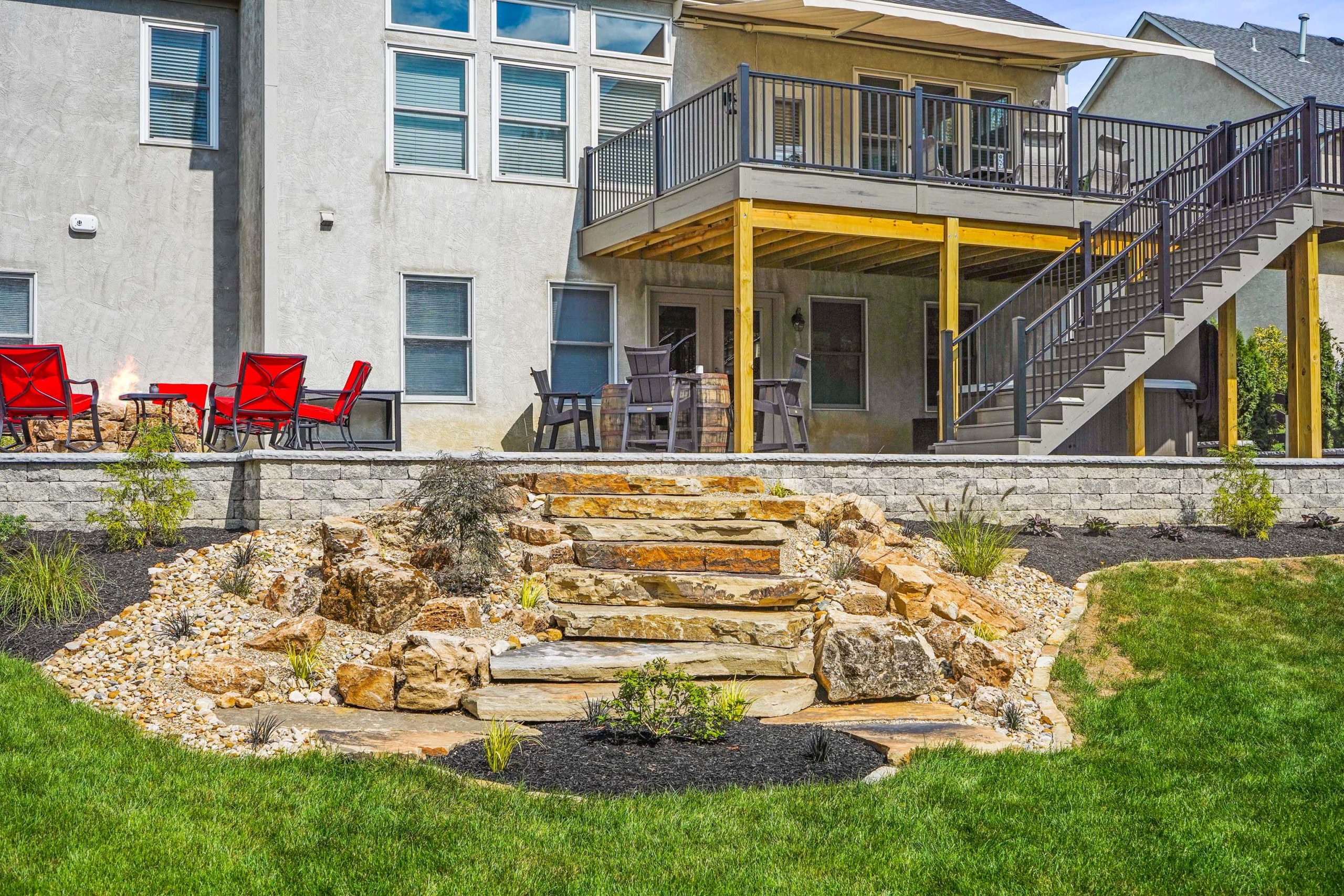 Full view of a storey house with patio upstairs, jacuzzi in shade downstairs, fireplace with red rocking chairs, stairs hardscape surrounded by shrubs and lawn.