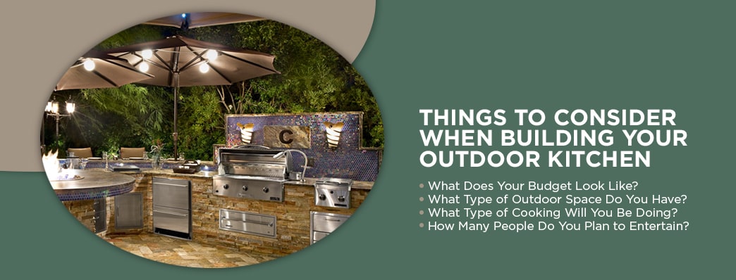 At dusk, an outdoor kitchen set with umbrellas over the counters with lights shinning down and illuminating the area, all cropped in an oval with text to the right that reads "Things to Consider When Building Your Outdoor Kitchen" followed by four bullet points which read "What Does Your Budget Look Like? What Type of Outdoor Space Do You Have? What Type of Cooking Will You Be Doing? How Many People Do You Plan to Entertain?"