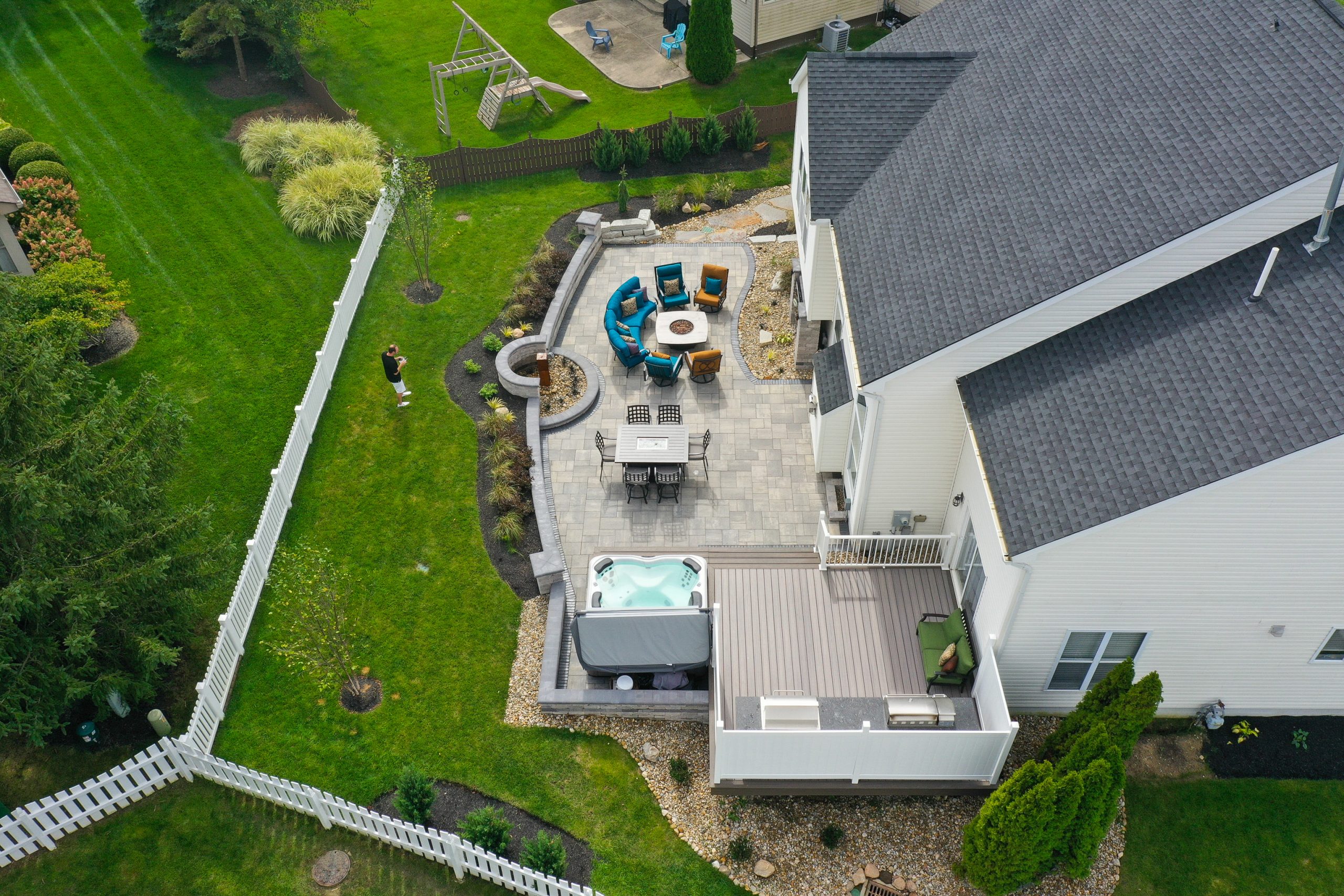 Top view of home backyard lawn within fence with a man, shrubs, lounge area, dining area and jacuzzi side-by-side a kitchen with hedges behind. Outside the fence are shrubs, trees and a playground on lawn.